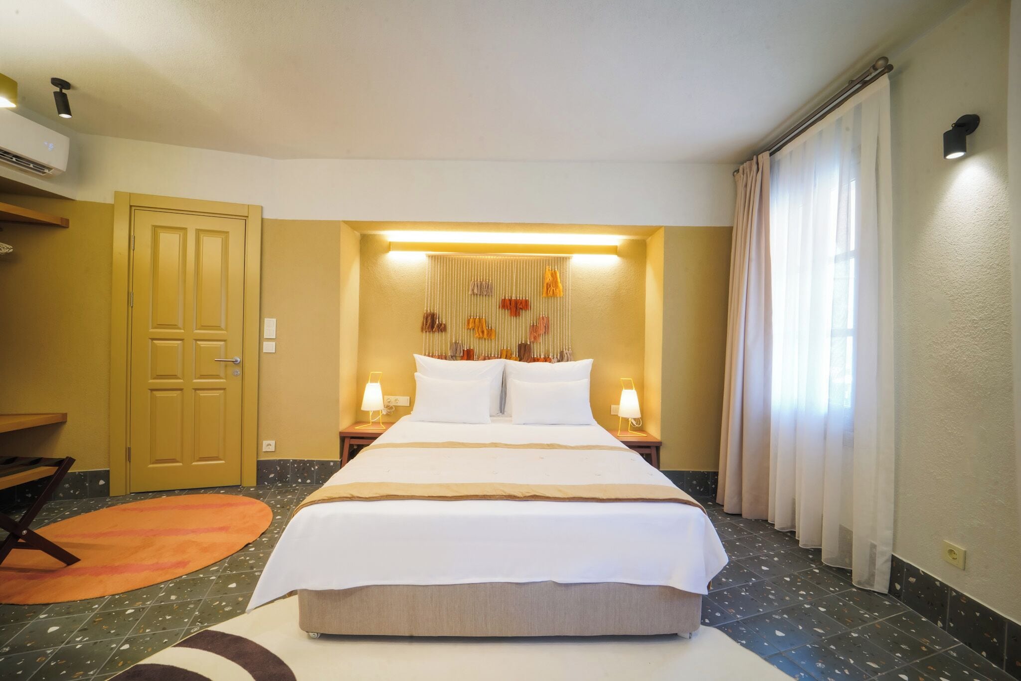 A hotel room with a large bed, a desk, and a chair. There is a window with curtains, a wall-mounted air conditioner, and a light fixture above the bed.