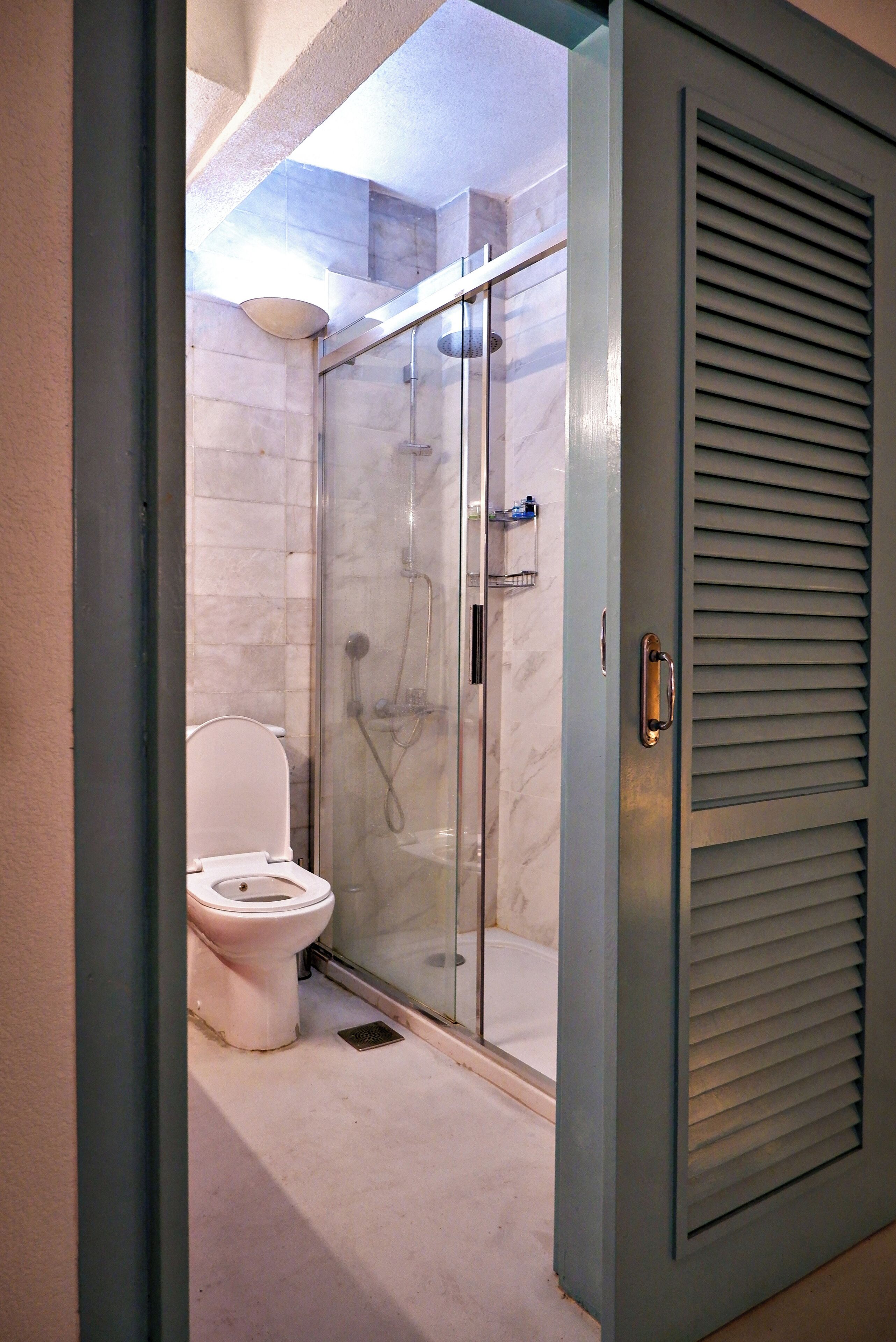 Une salle de bain comprenant des toilettes, une douche à parois de verre et une porte à lattes horizontales.
