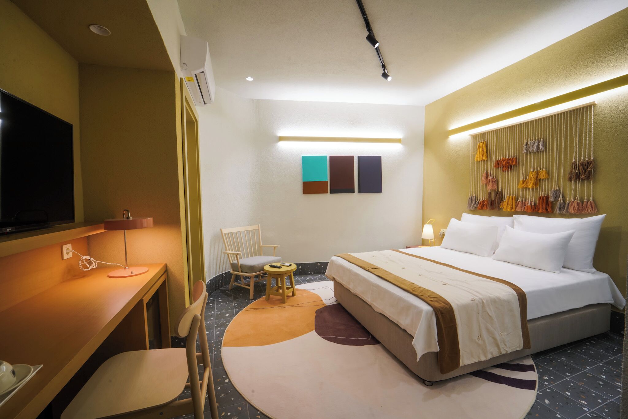 A hotel room with a bed, a desk, a chair, a TV, and a wall-mounted light fixture.