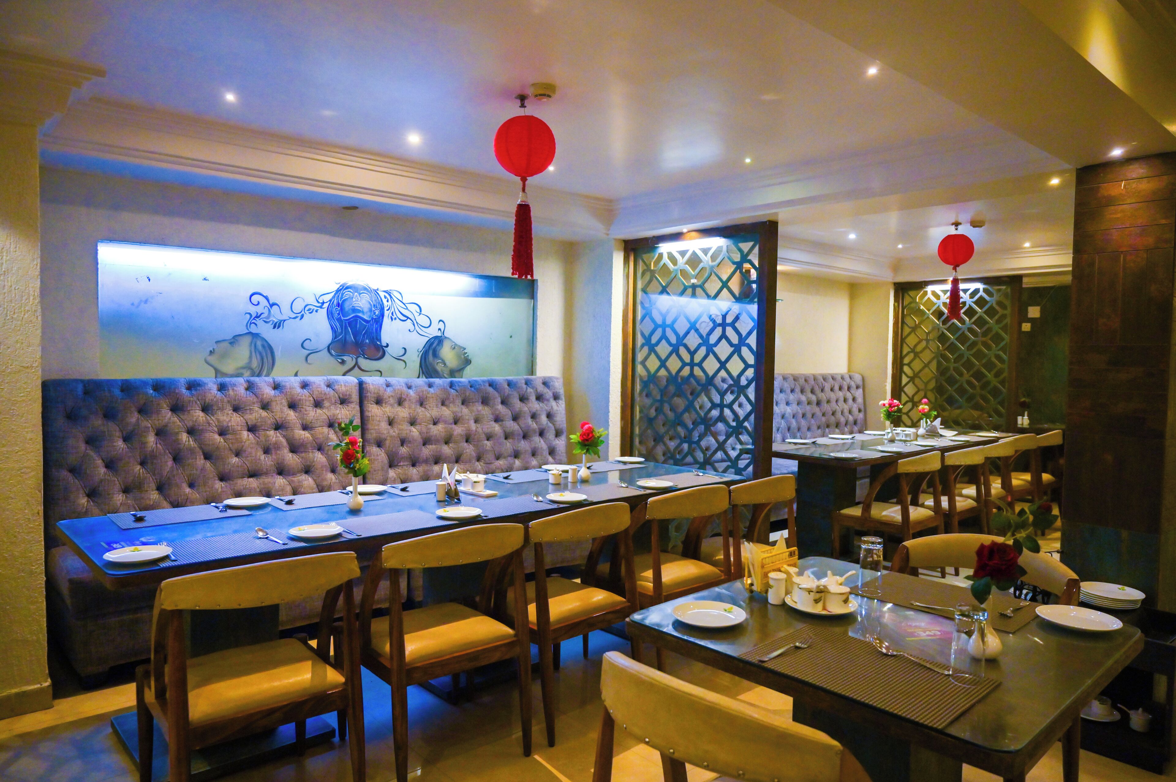 A dining area with a long table set for multiple people, cushioned seating, and decorative elements like a wall art piece and red lanterns.