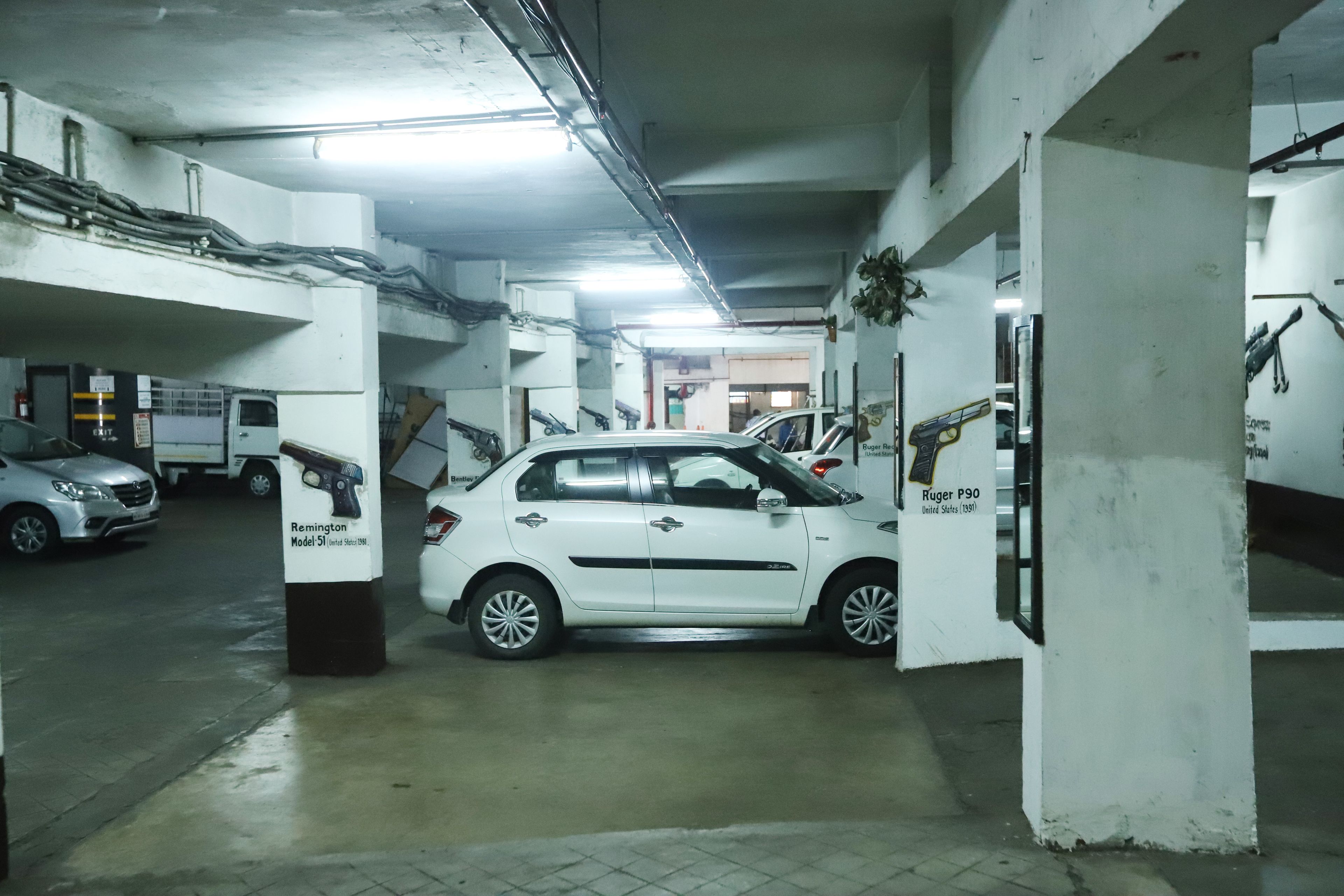Un parking aux murs blancs et au sol en béton. Plusieurs voitures sont garées, dont une berline blanche et un fourgon blanc.