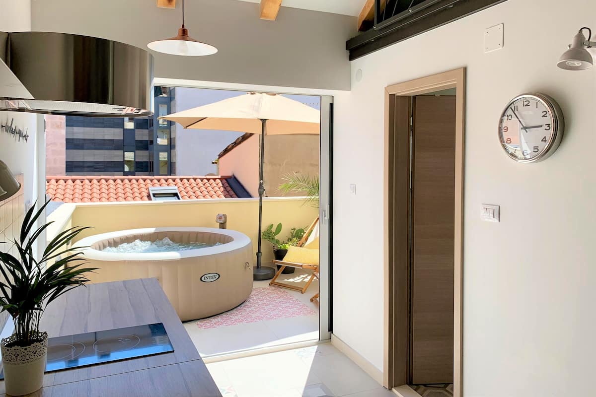 A modern bathroom with a bathtub, a sink, and a shower area. There is a clock on the wall and a view of the outside balcony.
