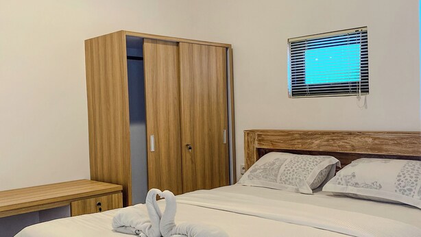 A neatly made bed with a towel sculpture, a wooden wardrobe, a bedside table, and a window with blinds.
