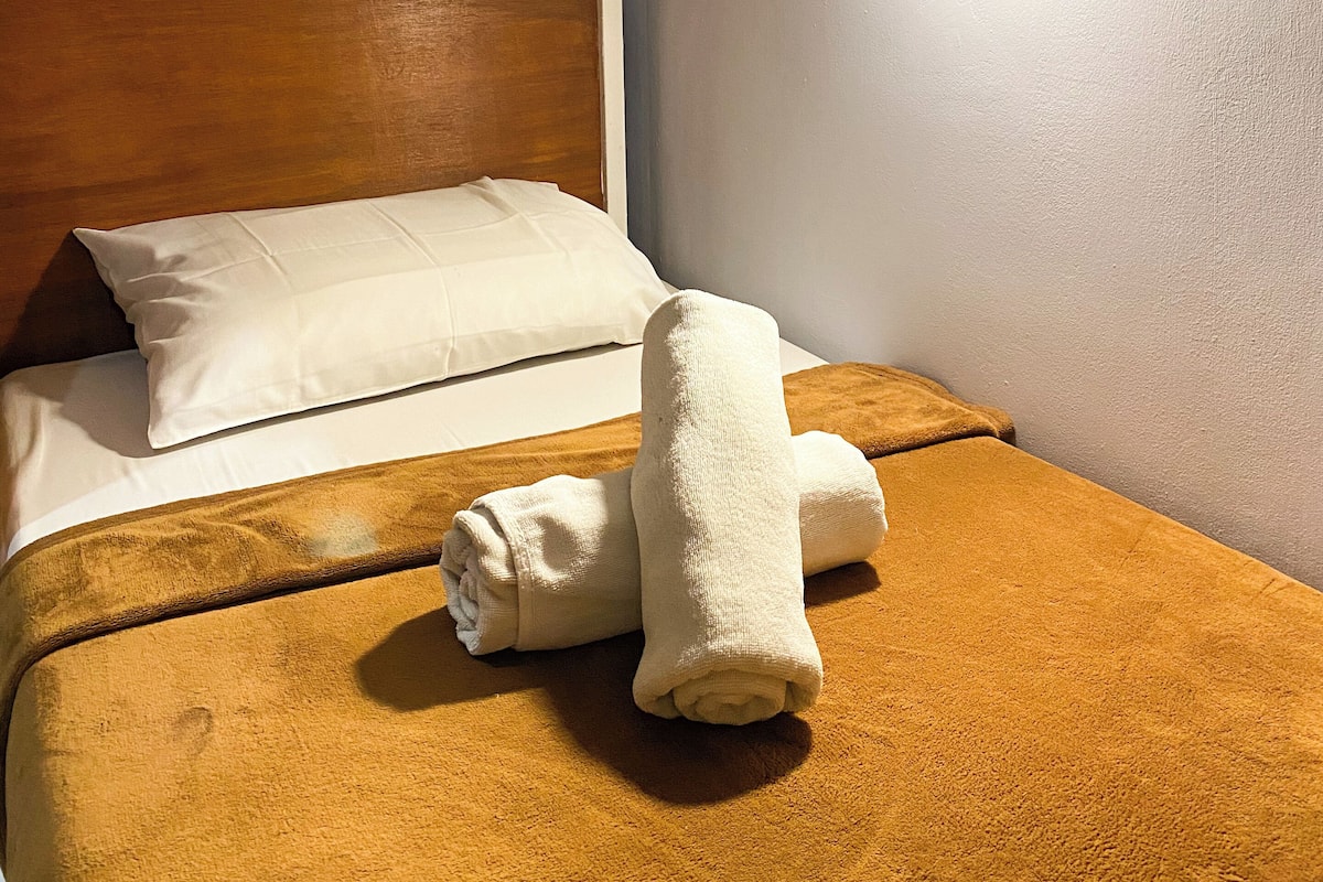 Una habitación de hotel con cabecera de madera, una cama individual con una manta marrón, un almohadón blanco y una toalla doblada sobre la cama.