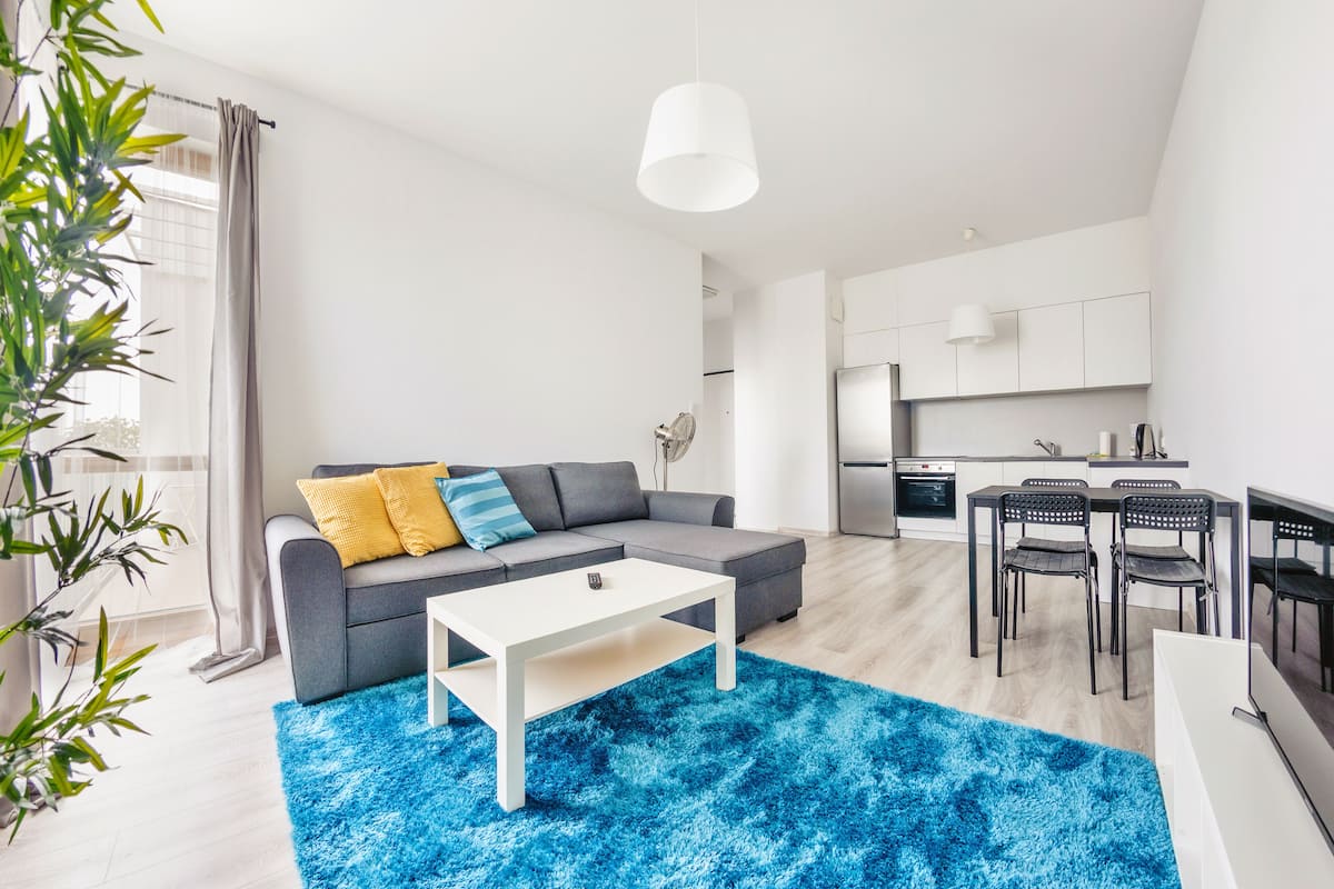 Una sala moderna con un sofá gris, una mesita de centro blanca y un tapete azul. Se observa una cocina con armarios blancos y una mesa de comedor con sillas.