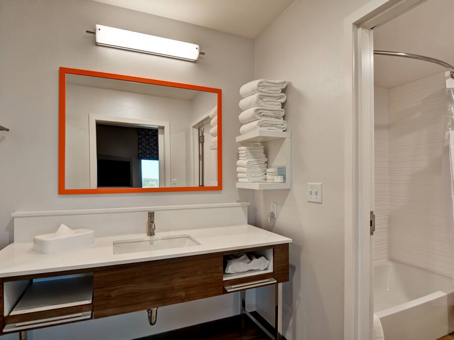 A modern bathroom with a white sink, wooden vanity, a large mirror with an orange frame, a towel rack, and a shower area.