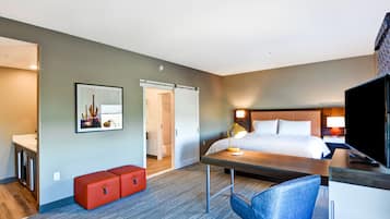 A hotel room with a bed, a desk, a chair, a TV, a red bench, and a door leading to a bathroom.