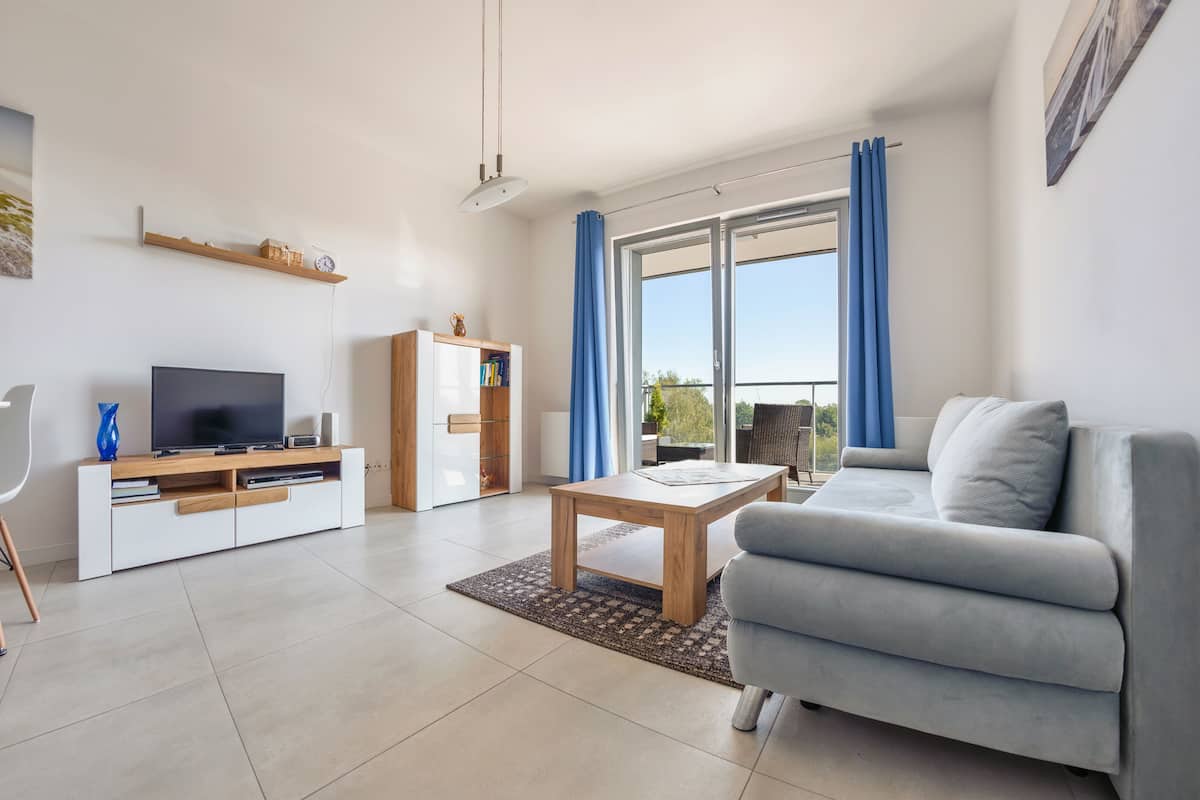 A modern living room with a flat-screen TV, a sofa, a coffee table, and a sliding glass door leading to a balcony.
