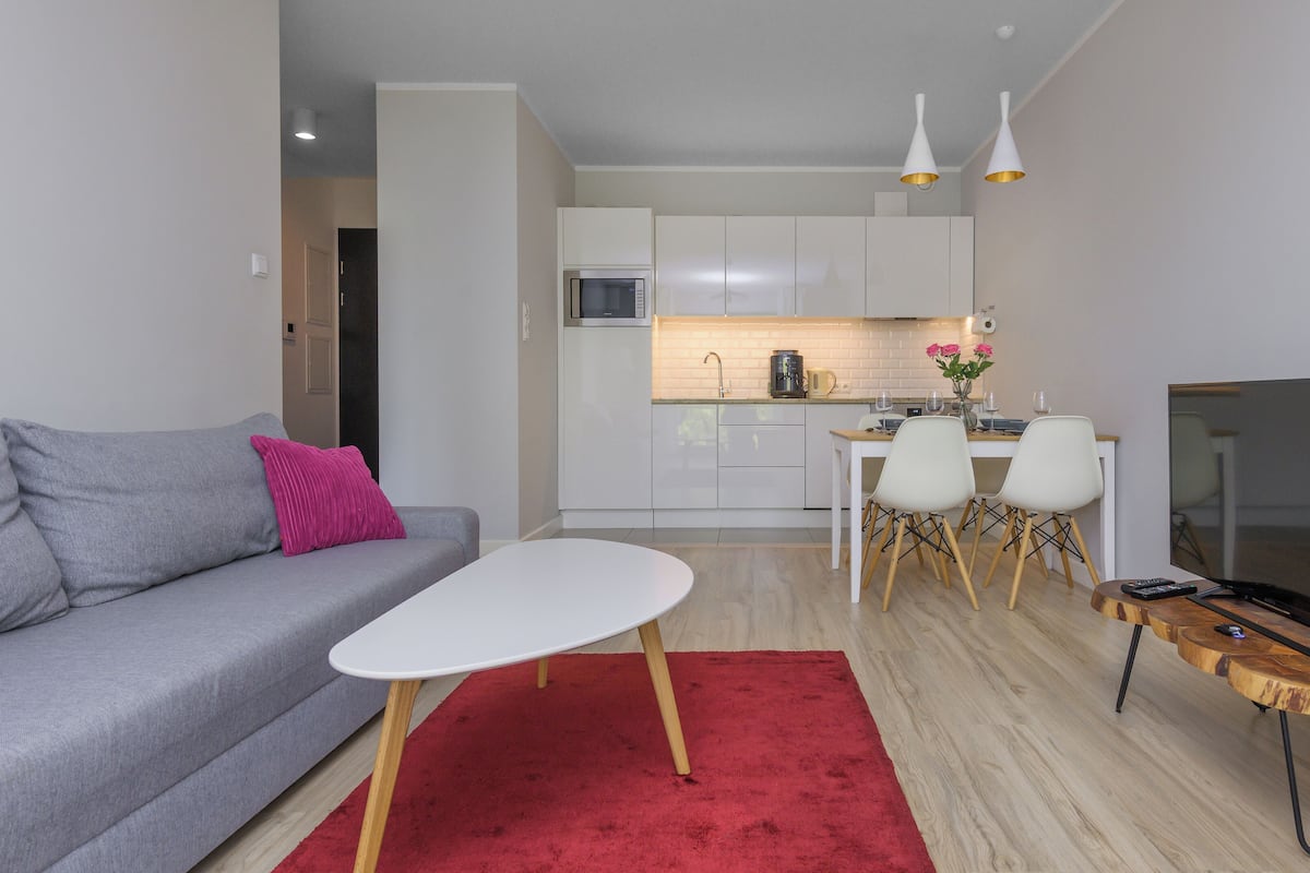 A modern living room with a grey sofa, a white coffee table, and a red rug. There is a kitchen area with white cabinets and a dining table with chairs. A TV is on a wooden stand, and there are two pendant lights above the kitchen.