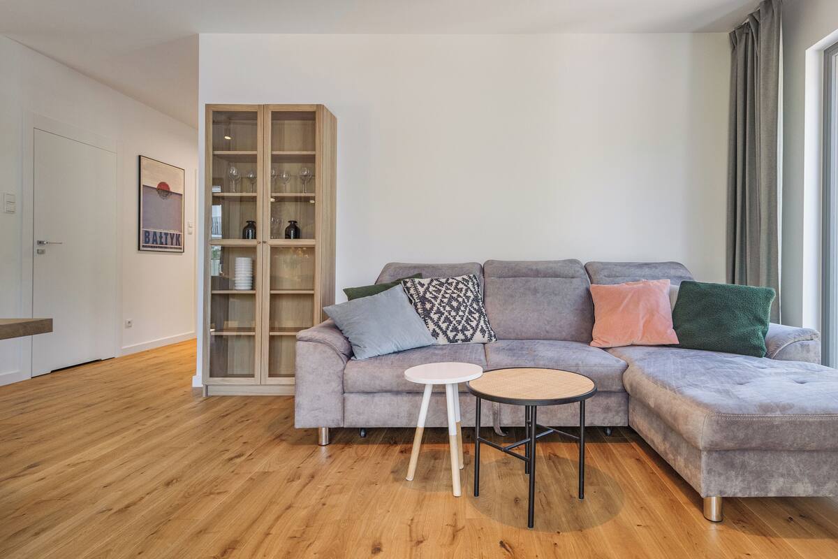 A modern living room with a grey sectional sofa, a small round table, and a glass cabinet with wine glasses.