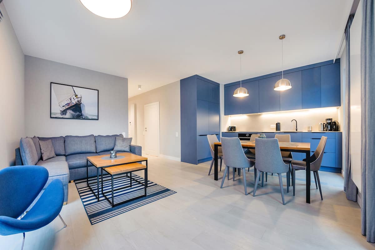 A modern living room with a sofa, a dining table, and chairs. There is a kitchen area with cabinets and a countertop.