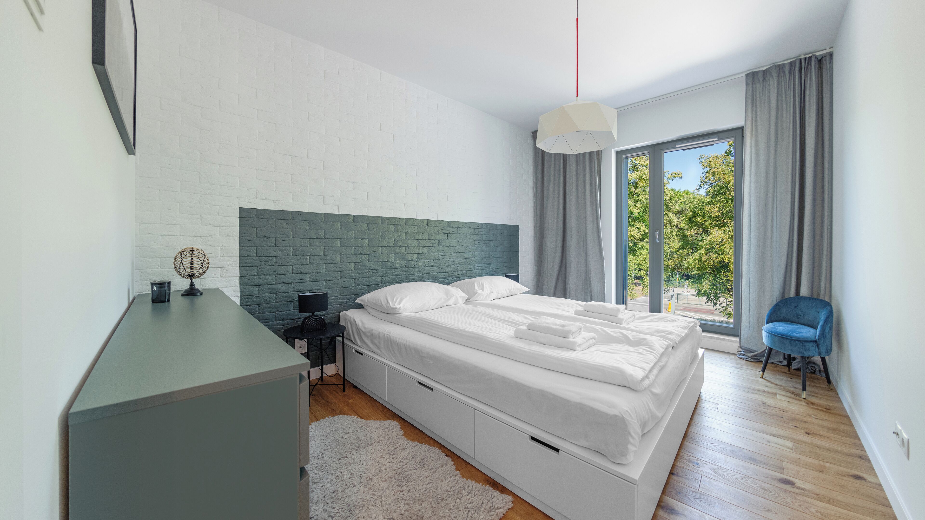 Ein modernes Schlafzimmer mit einer weißen Ziegelwand, einem grünen Kopfteil, einem großen Fenster mit Blick auf Bäume, einem Nachttisch mit Lampe und einem blauen Sessel.