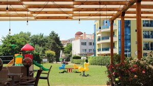 An outdoor patio area with wooden flooring, wicker furniture, and a children's play structure. - Aparthotel Marina Holiday Club & Spa (Pomorie)