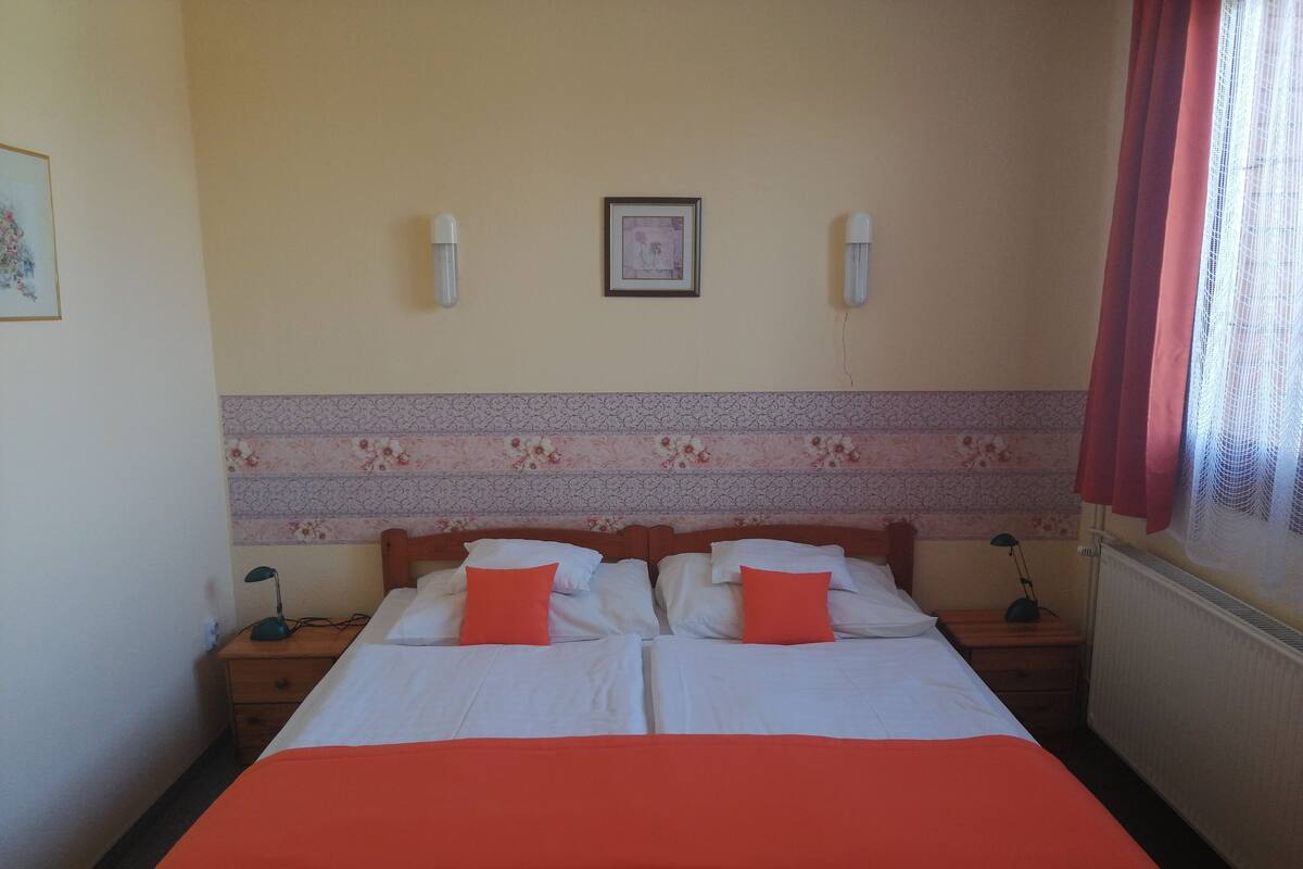 A double bed with an orange bedspread, two bedside tables with lamps, a framed picture on the wall, and a window with a red curtain.
