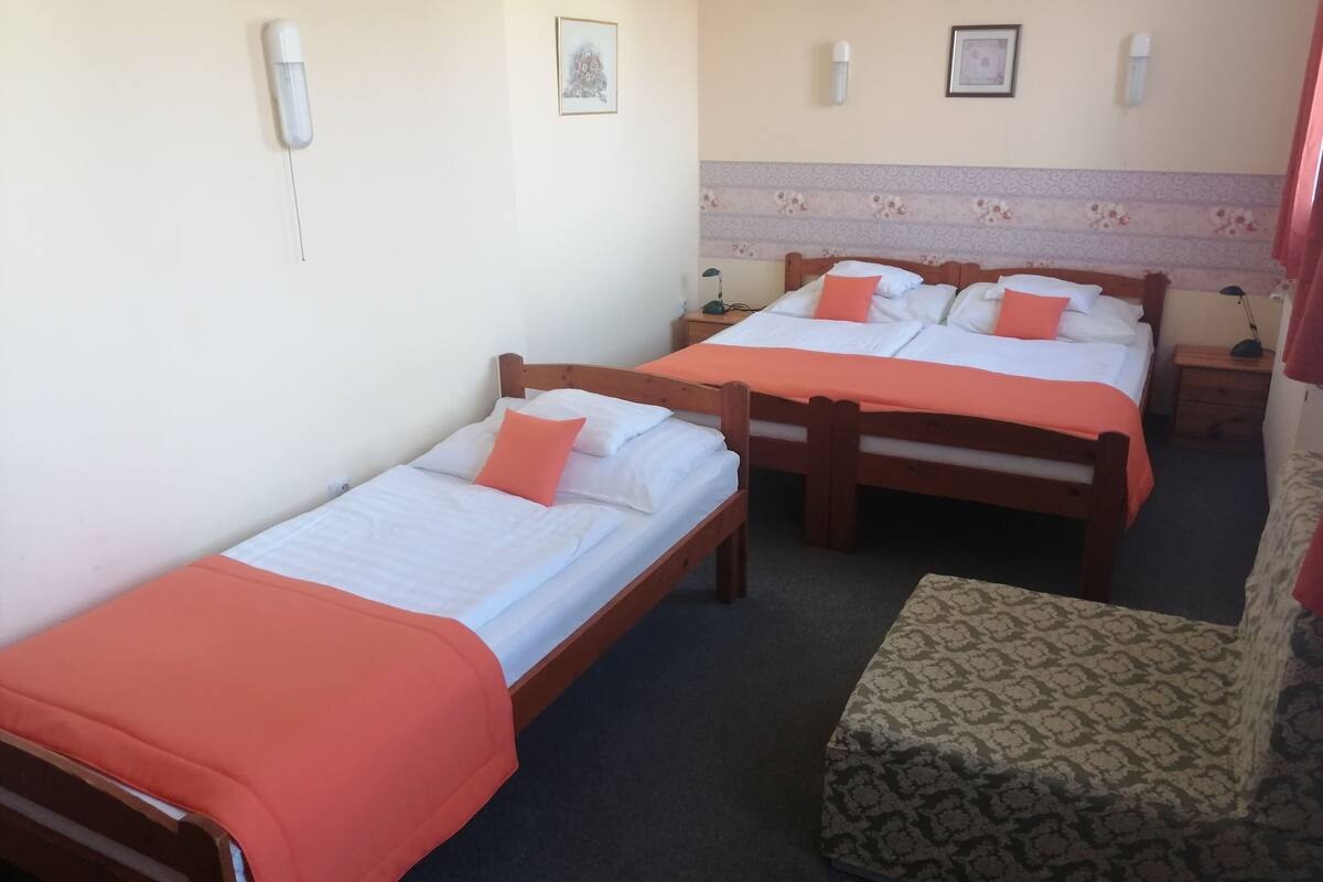 A hotel room with two wooden beds, each with white linens and orange pillows. There is a patterned armchair and a small table with a lamp.