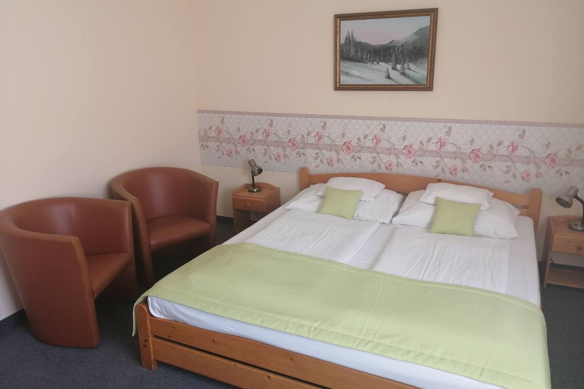 A hotel room with a wooden bed, two chairs, a nightstand with a lamp, and a floral wallpaper border.