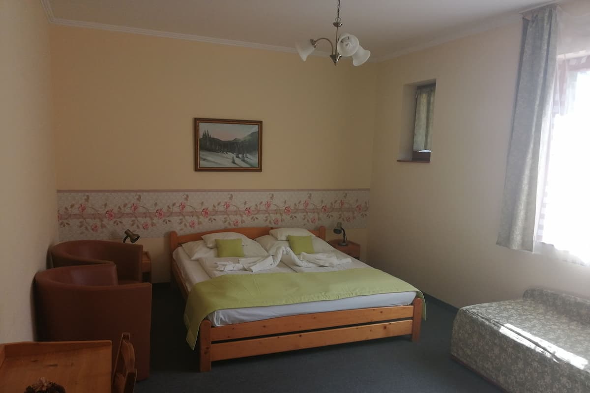 A bedroom with a wooden bed, a desk, a chair, a small table with a plant, and a window with curtains.