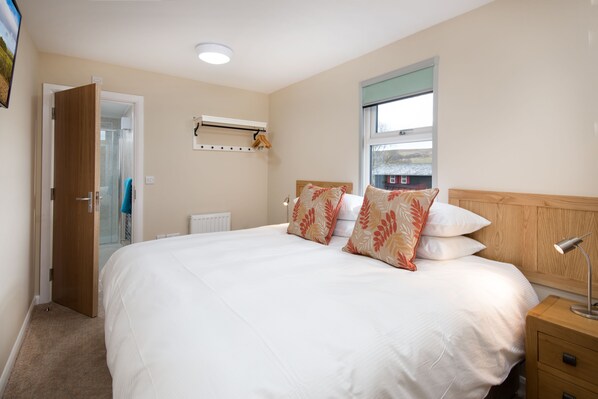 A hotel room with a large bed, a wooden headboard, a bedside table with a lamp, a window with a view, and a door leading to a bathroom.