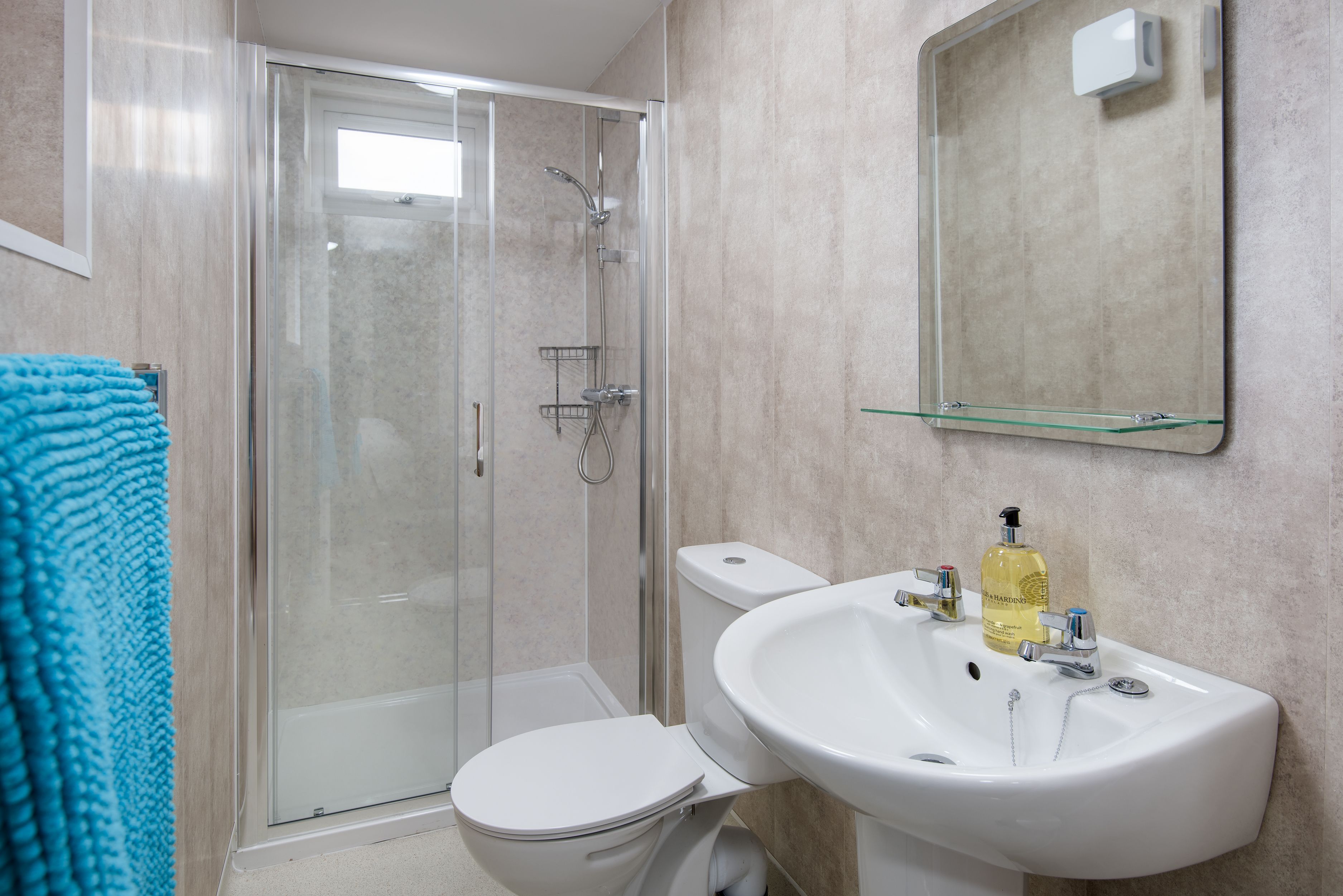 A bathroom with a glass shower enclosure, a white toilet, a sink with a mirror above it, and a blue towel on the floor.
