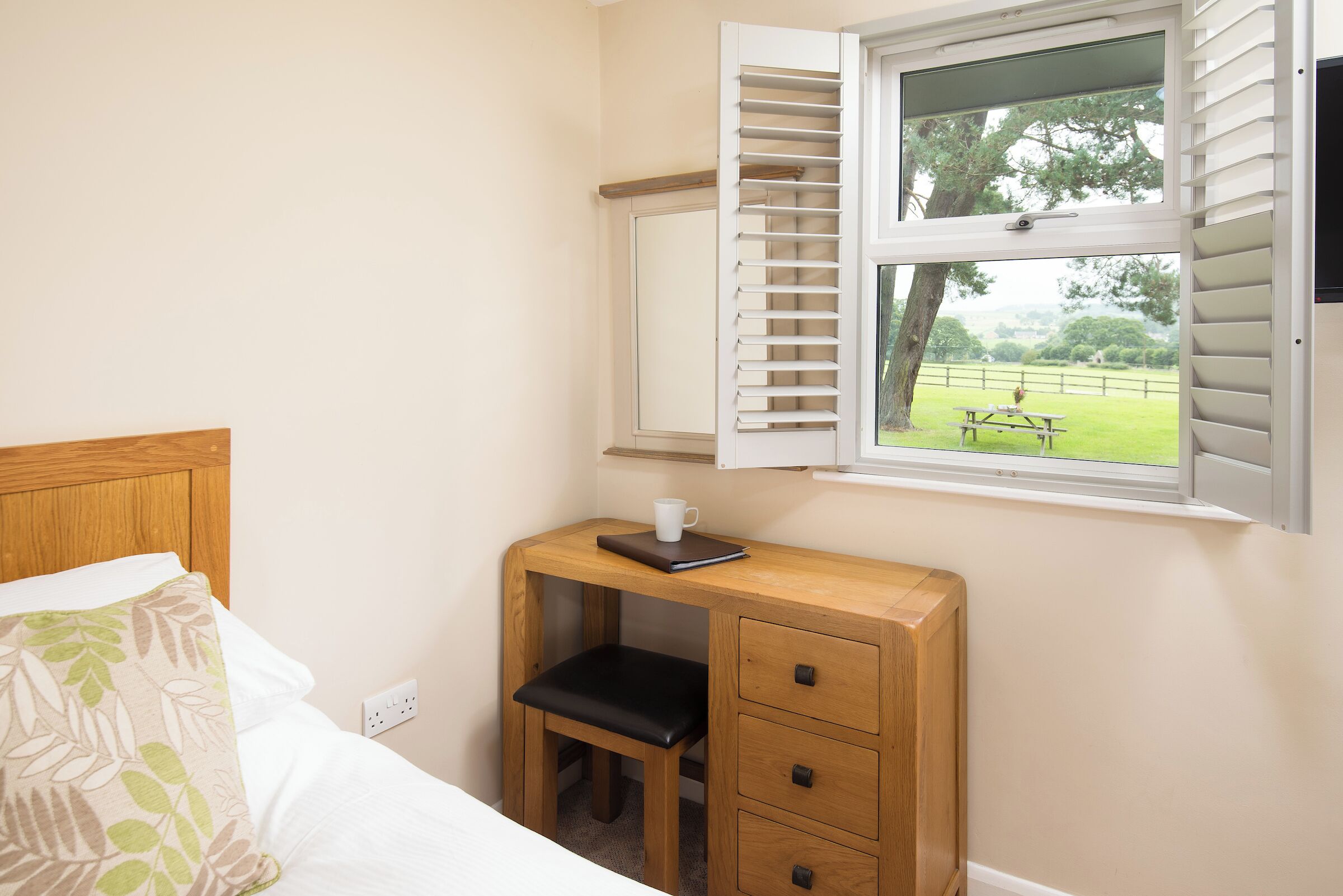 Een slaapkamer met een bed, een houten bureau met stoel, een raam met luiken en uitzicht op een grasveld.