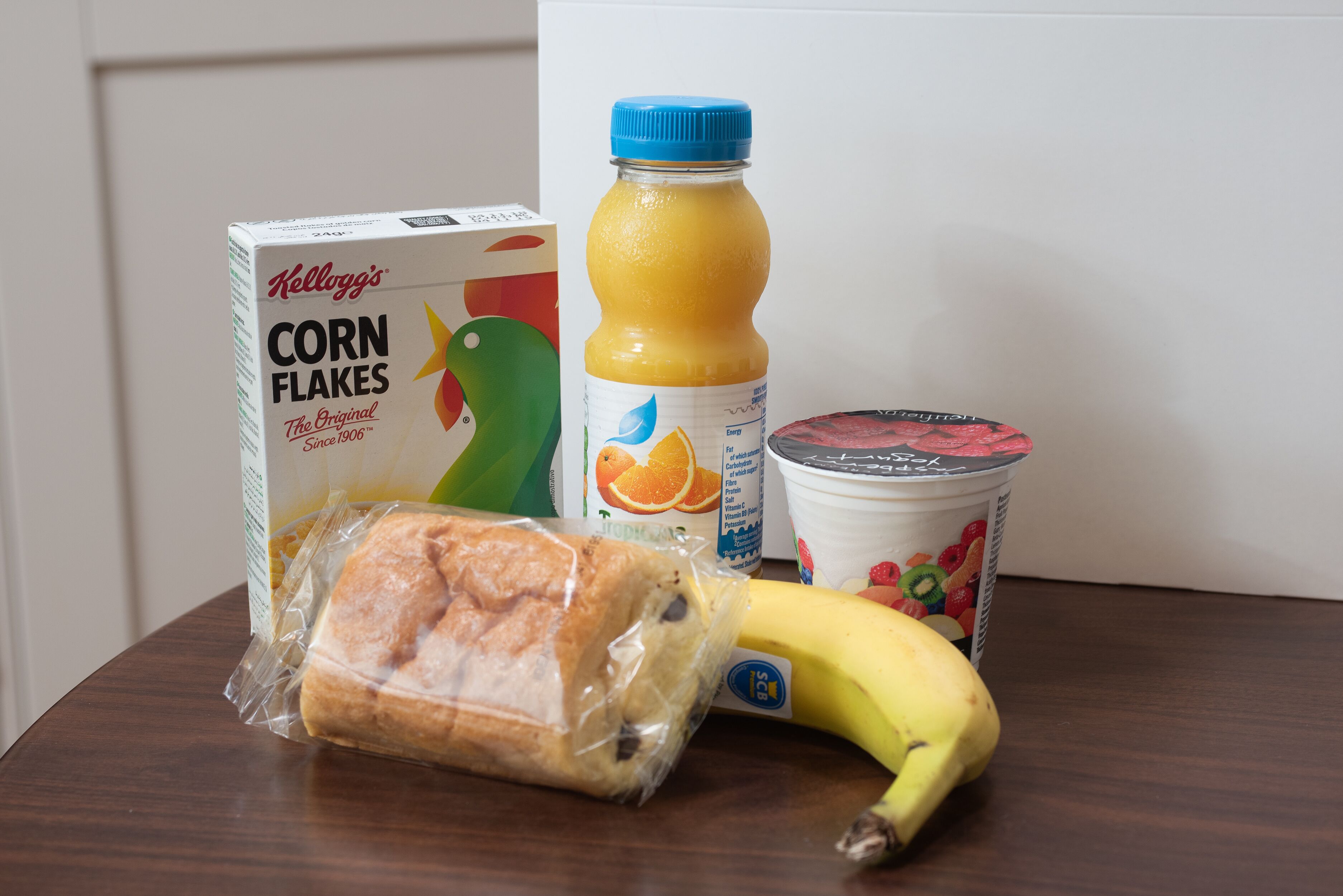 Una scatola di Kellogg's Corn Flakes, una bottiglia di succo d'arancia, una banana, un vasetto di yogurt e una pagnotta di pane su un tavolo di legno.