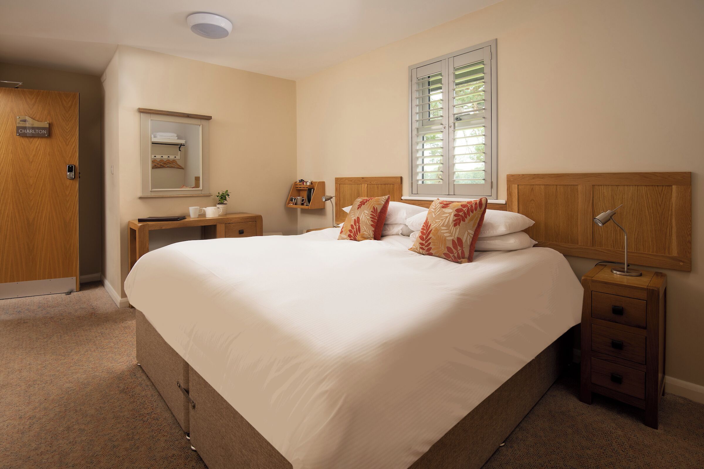 A hotel room with a large bed, a bedside table, a nightstand, a mirror, and a window with blinds.