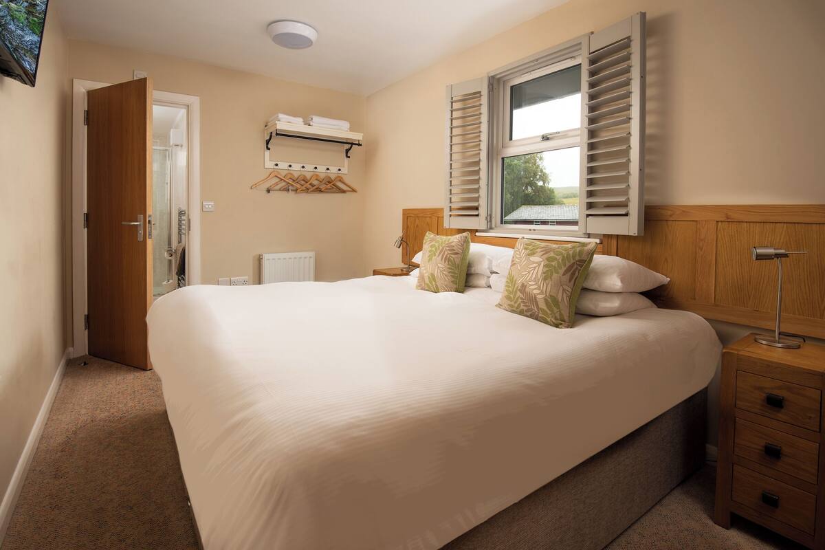 A bedroom with a large bed, a bedside table, a nightstand, a window with shutters, a radiator, and a wall-mounted TV.