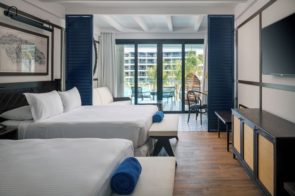 Habitación de hotel con dos camas, televisión, balcón con vistas a la piscina y edificios, y zona de comedor con sillas.