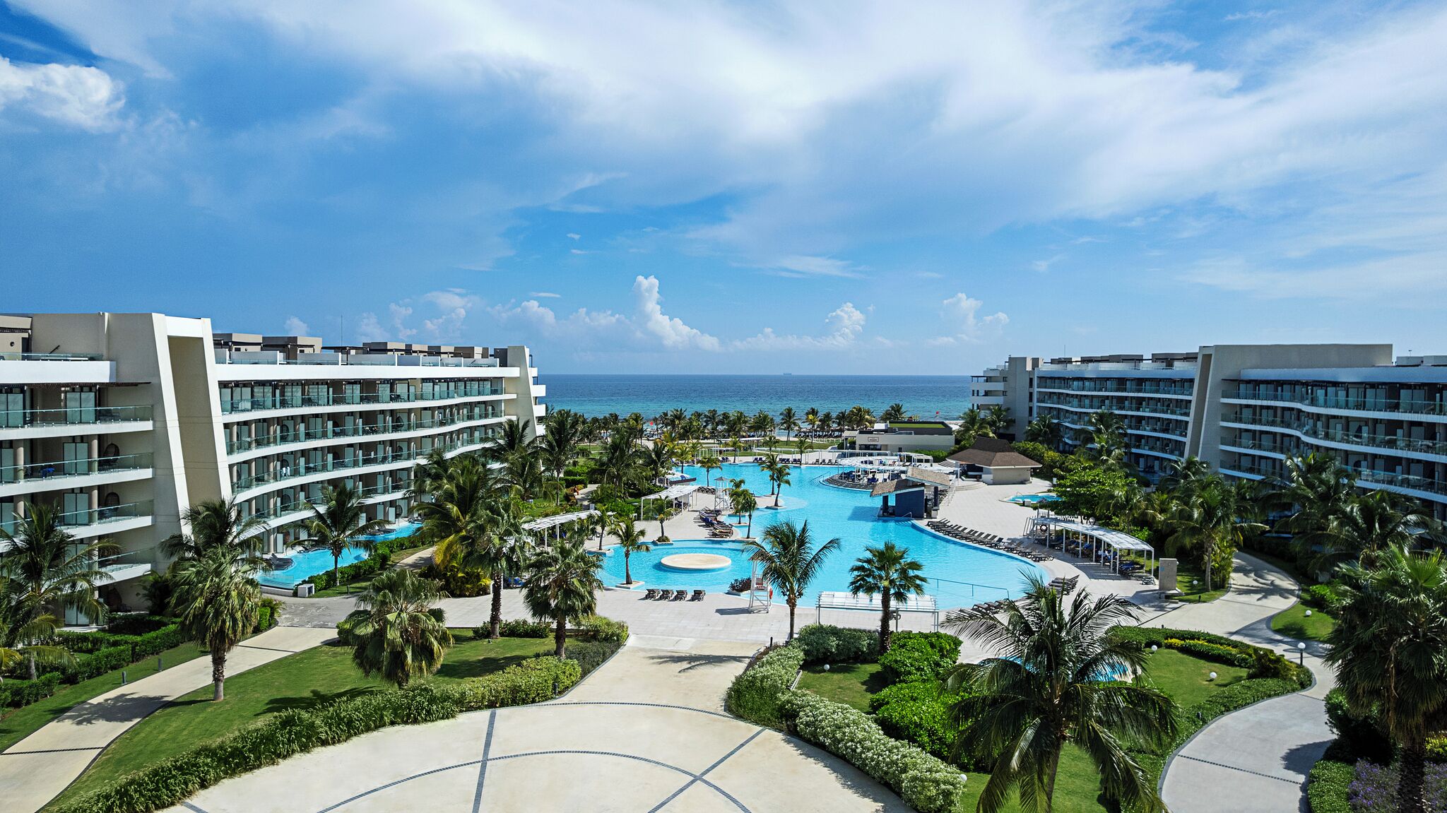 A resort with a central pool, multiple buildings, and a clear view of the ocean.