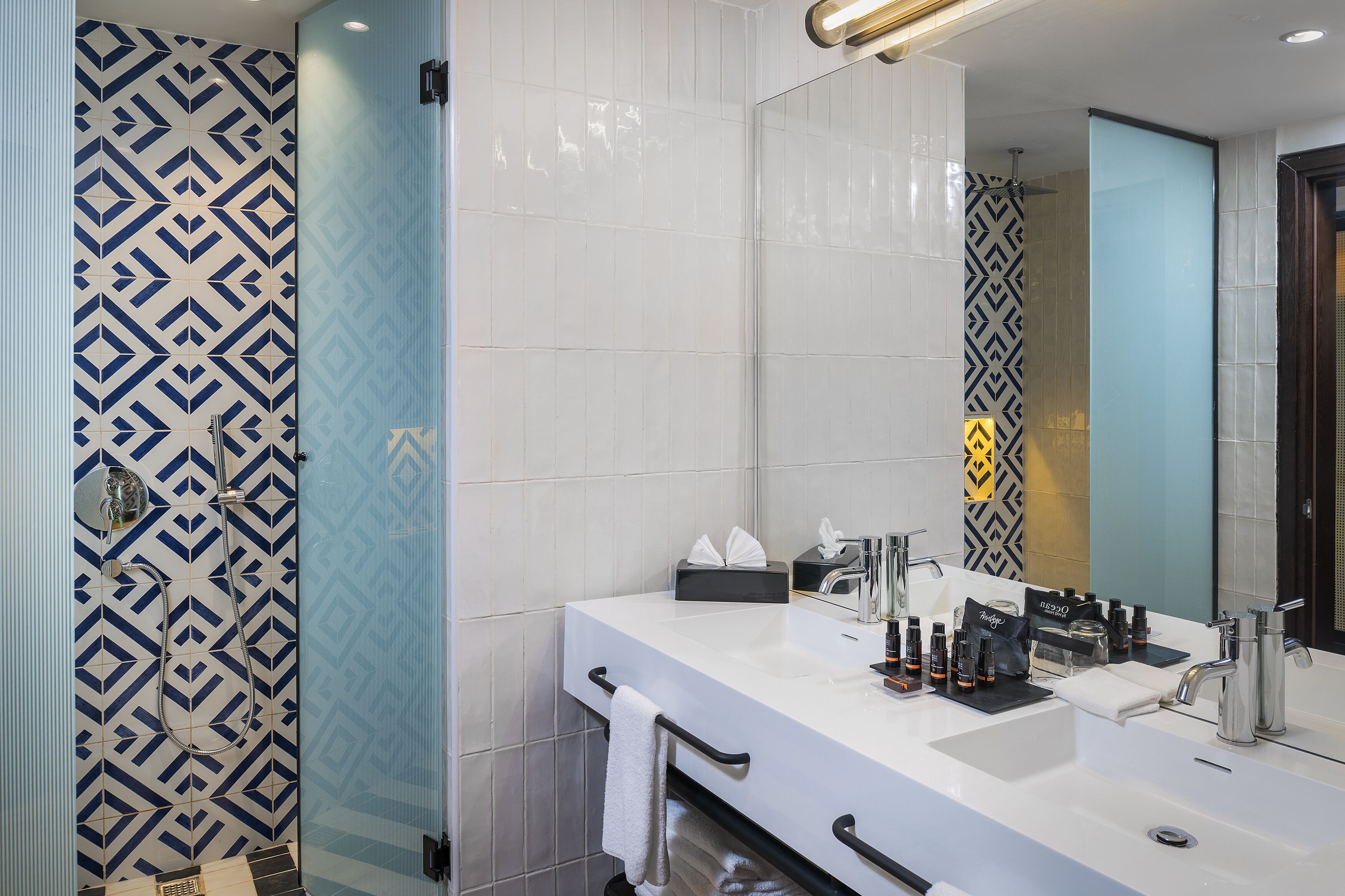 Un baño moderno con un lavamanos blanco, un espejo grande y una ducha con un patrón geométrico de azulejos.