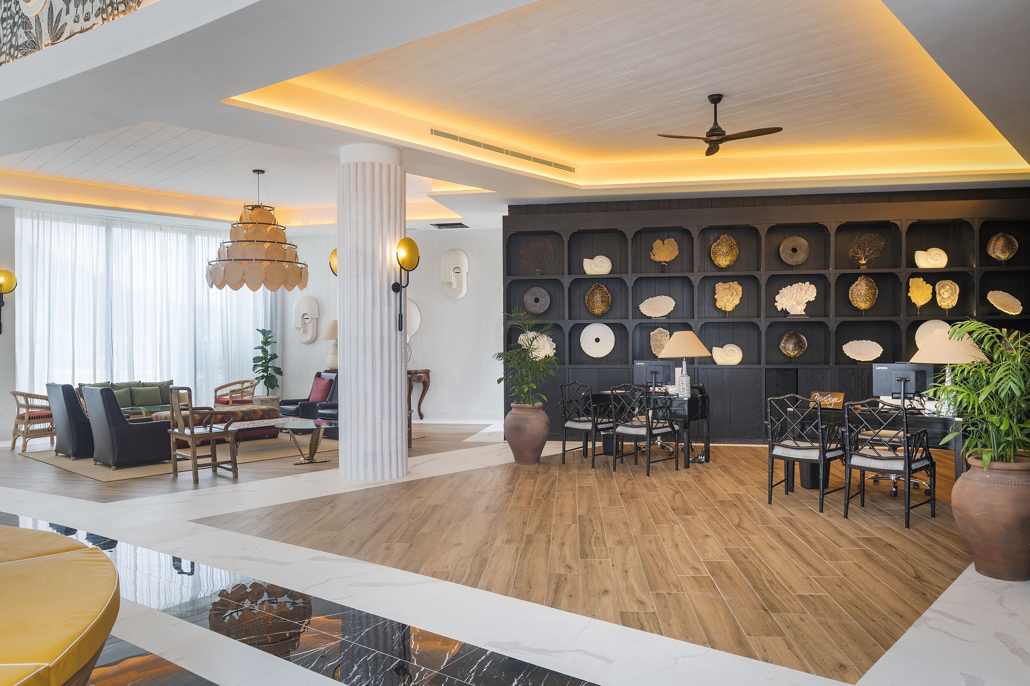 Un hall d’entrée spacieux doté d’un plancher en bois, un espace de détente central avec des fauteuils et un canapé, un ventilateur de plafond et un mur orné d’assiettes décoratives.
