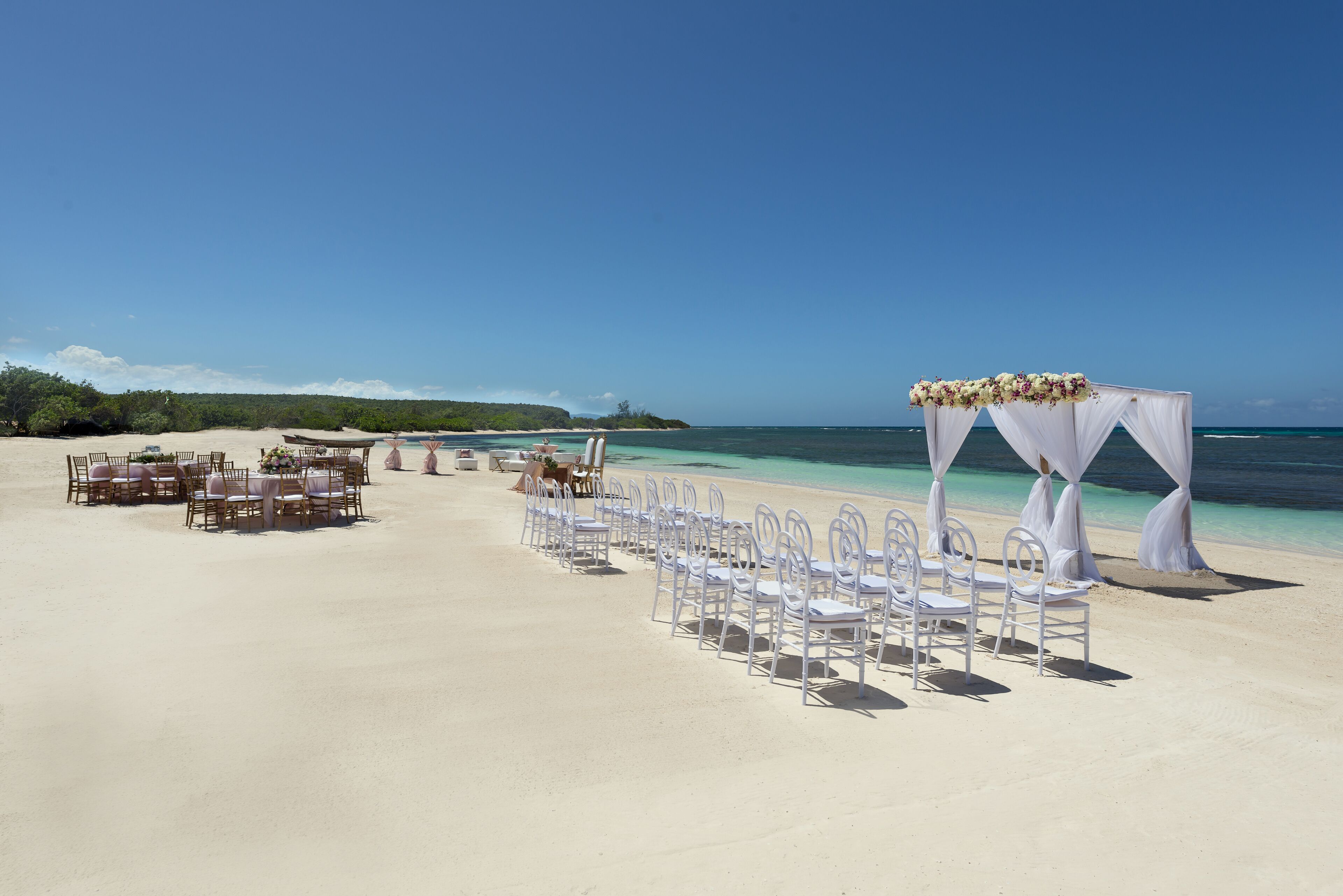 Une cérémonie de mariage organisée sur la plage, avec des chaises et des tables blanches, une arche décorée de fleurs et une vue sur l’océan.