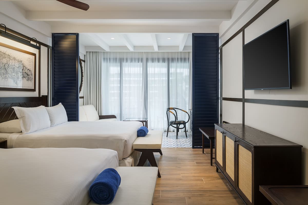 Habitación de hotel con dos camas, un escritorio, una silla, un televisor y un ventanal con cortinas.