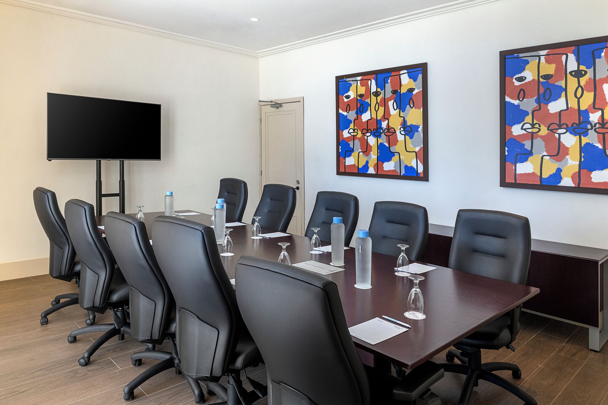 A conference room with a long table, black chairs, a flat-screen TV, and abstract paintings on the walls.