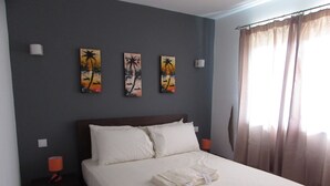 Una cama bien hecha con sábanas blancas, un cabecero de madera oscura y tres cuadros colgados en la pared que representan escenas tropicales.