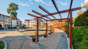 A covered outdoor area with a tiled walkway, wooden pergola, potted plants, and residential buildings in the background. - Apartamenty Sun & Snow Neptun Park (Gdansk)