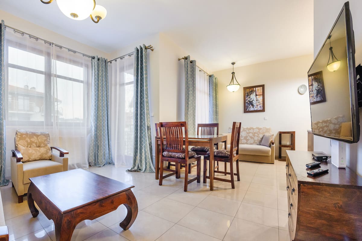 Una sala de estar con un sofá, una mesa de centro de madera, una mesa de comedor con sillas, un televisor y cortinas.