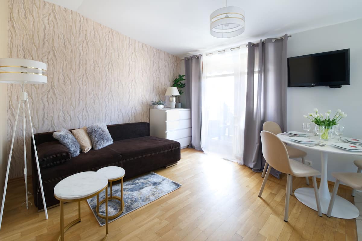 Una sala moderna con un sofá marrón oscuro, una mesa de centro redonda, una mesa de comedor preparada para dos personas y un televisor de pantalla plana montado en la pared.