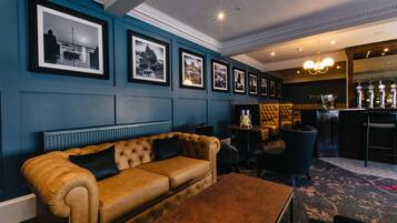 A lounge area with a brown leather sofa, framed black and white photographs on the wall, a bar with bottles, and a chandelier.