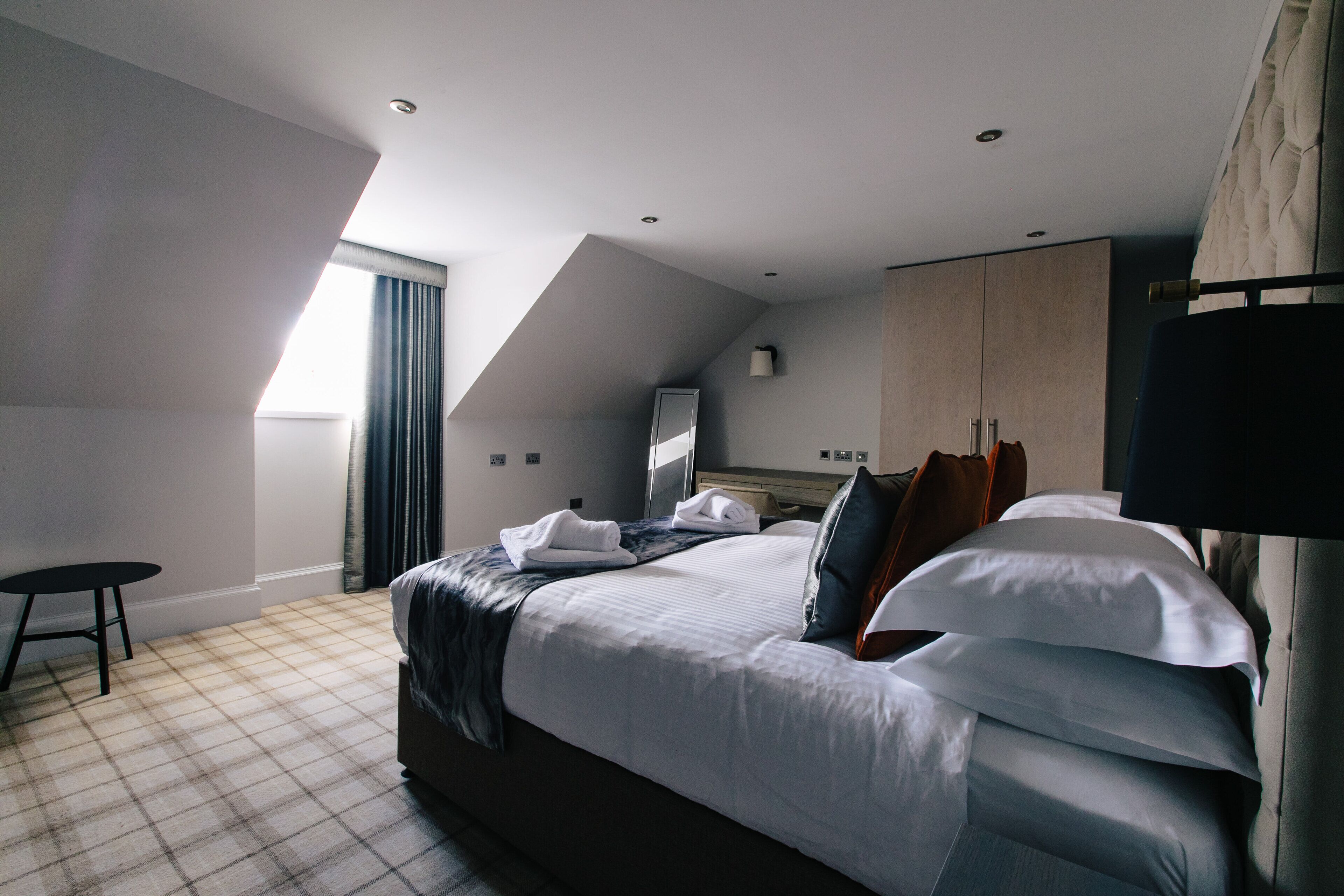 A modern hotel room with a large bed, a small table, and a view of the city through a window.