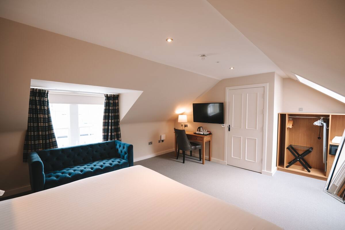 A hotel room with a large bed, a blue sofa, a desk with a chair, a TV, and a shower area.