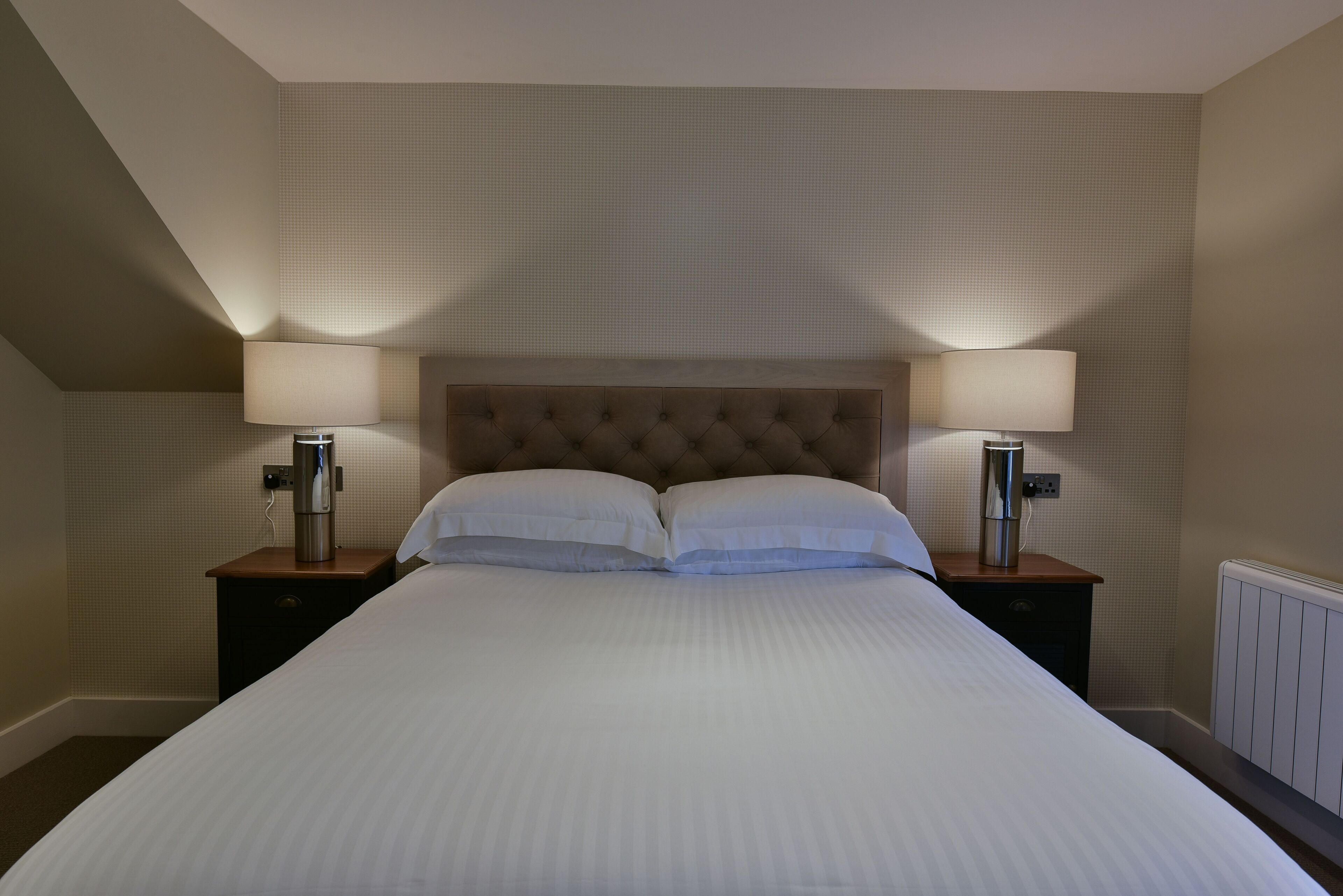 Una cama bien hecha con dos lámparas de noche y un calefactor en la pared.