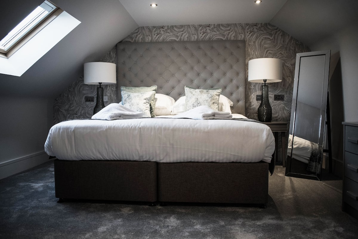 A bedroom with a large bed, two bedside lamps, a mirror, and a skylight.