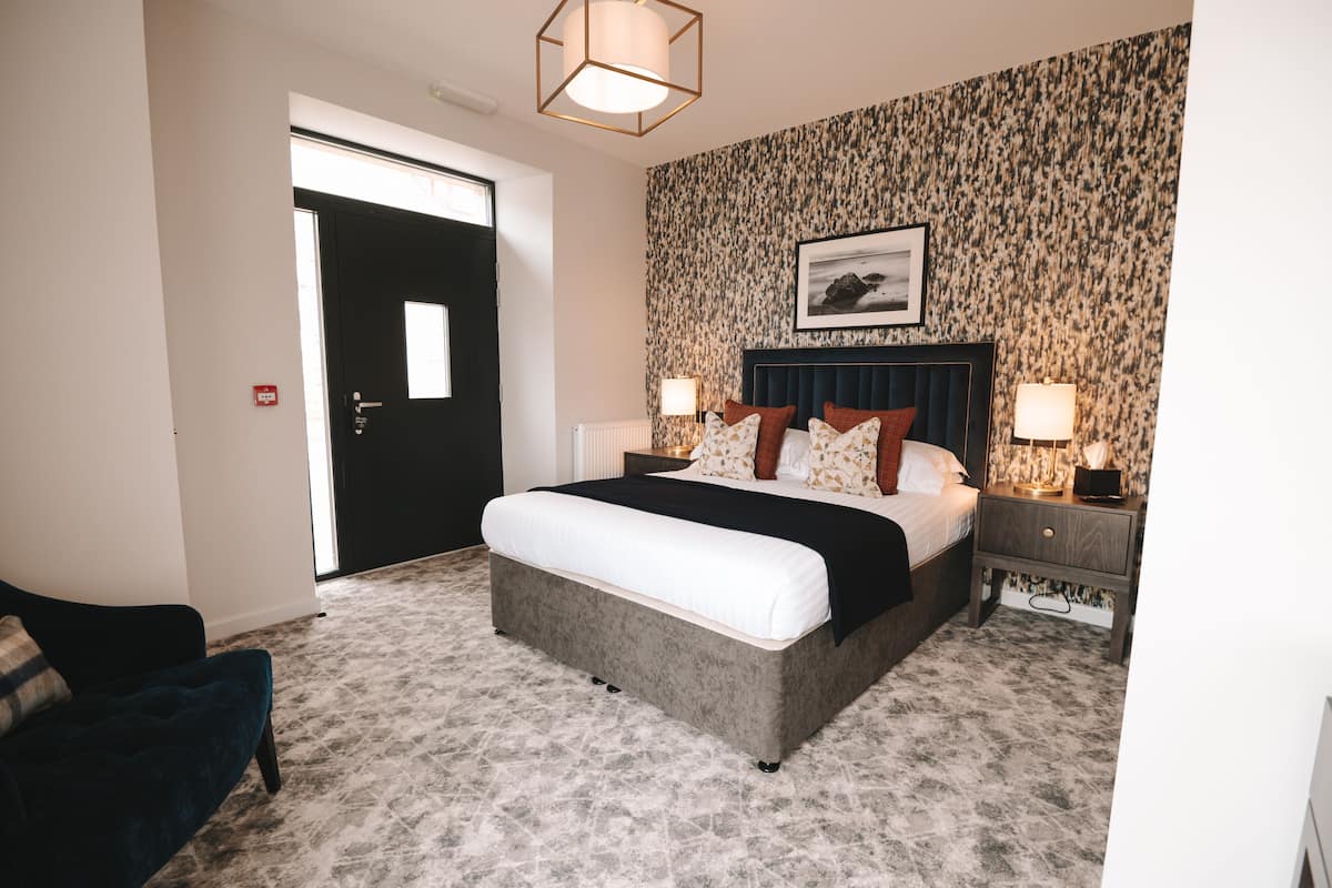A bedroom with a large bed, two bedside tables with lamps, a dark headboard, and a patterned wallpaper.