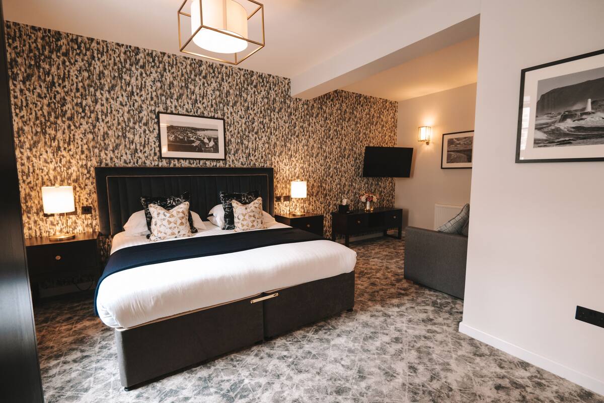 A bedroom with a large bed, a headboard, two bedside tables with lamps, a grey sofa, and a wall-mounted TV.