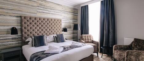 A modern hotel room with a tufted headboard, a large bed, two armchairs, a small table, and a view of the outdoors through a window with curtains.