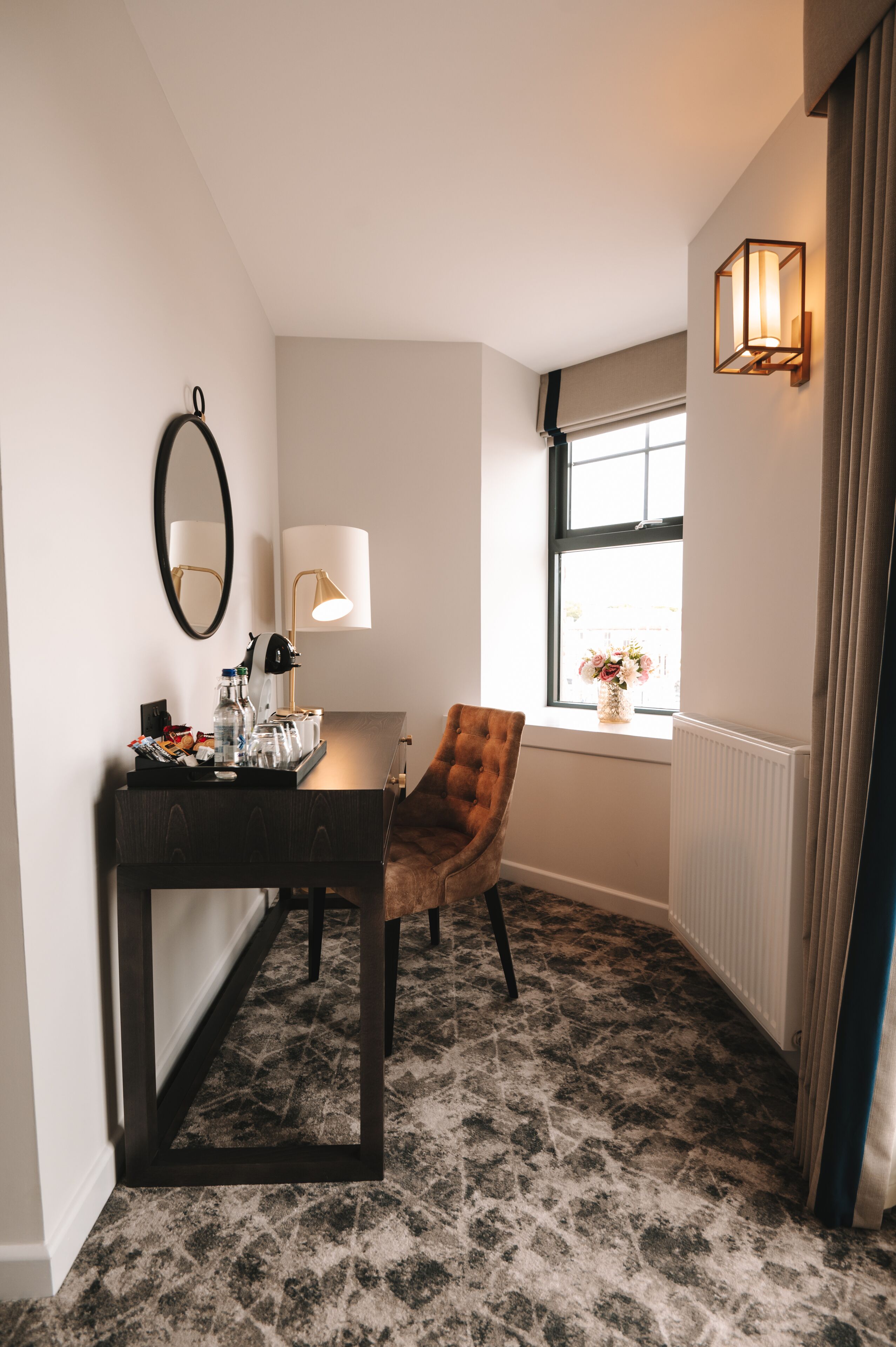 Une chambre d’hôtel avec un bureau en bois foncé, une chaise marron, un miroir, une lampe et une fenêtre donnant sur des fleurs.