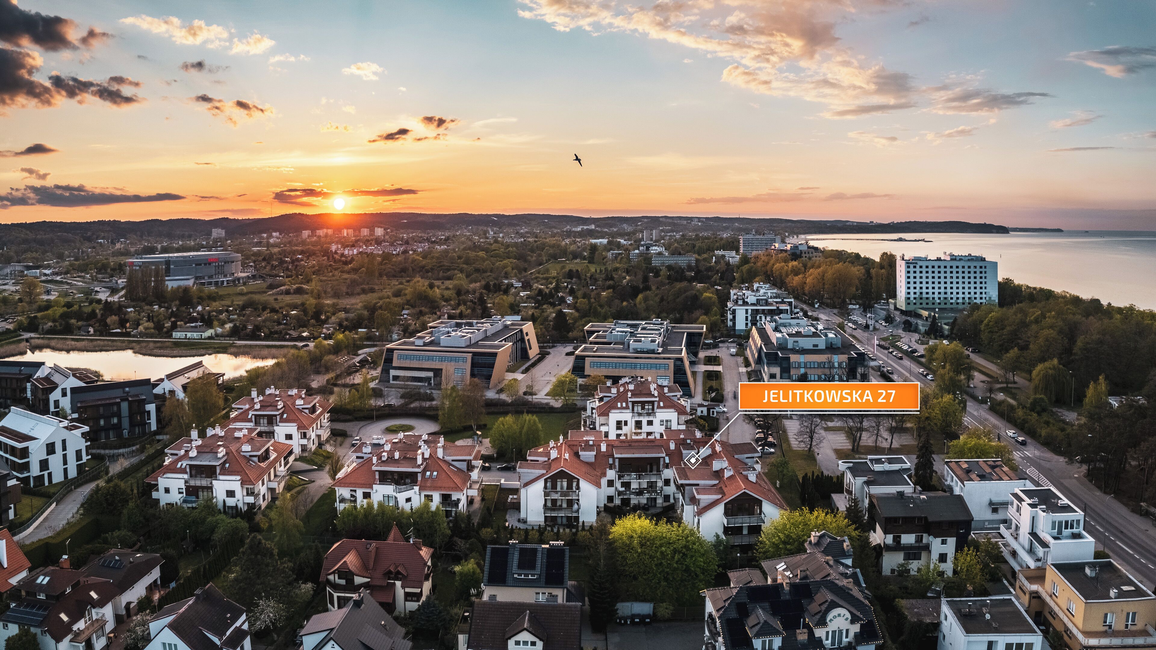 Luftaufnahme von Jelitkowska 27, mit einer Mischung aus Wohn- und Geschäftsgebäuden, einem Gewässer und einem Sonnenuntergang im Hintergrund.