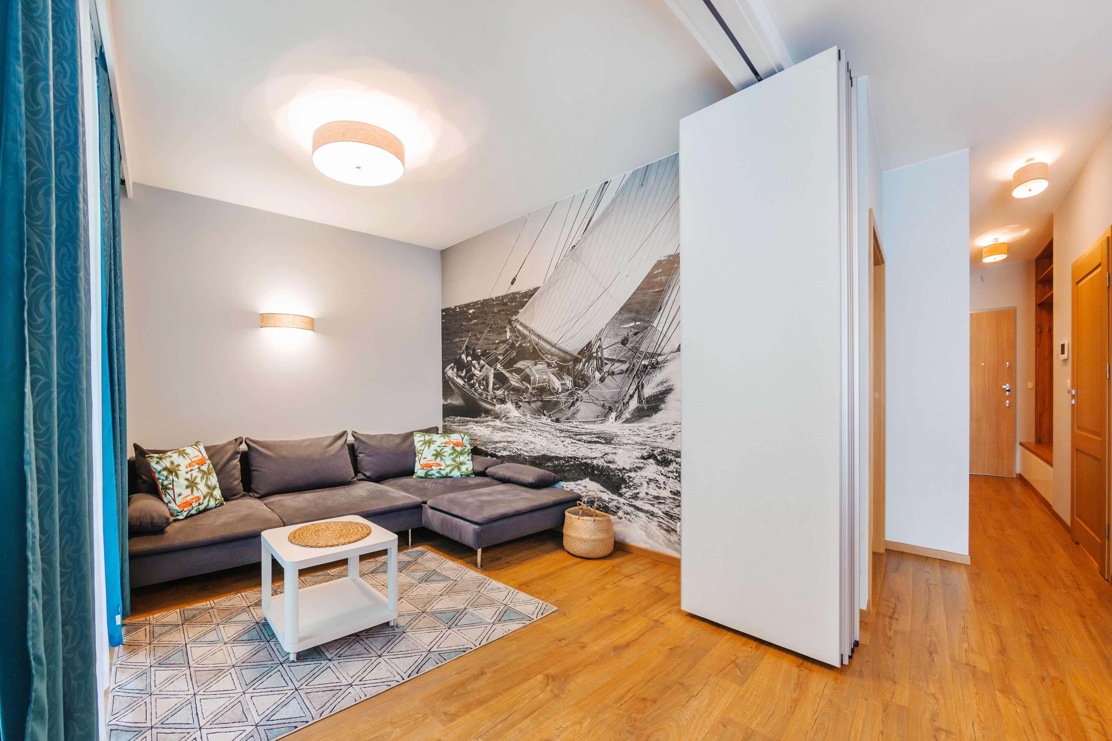 Een moderne woonkamer met een hoekbank, een klein wit bijzettafeltje, een kamerkleed met dessin en een groot zwart-wit muurschildering.