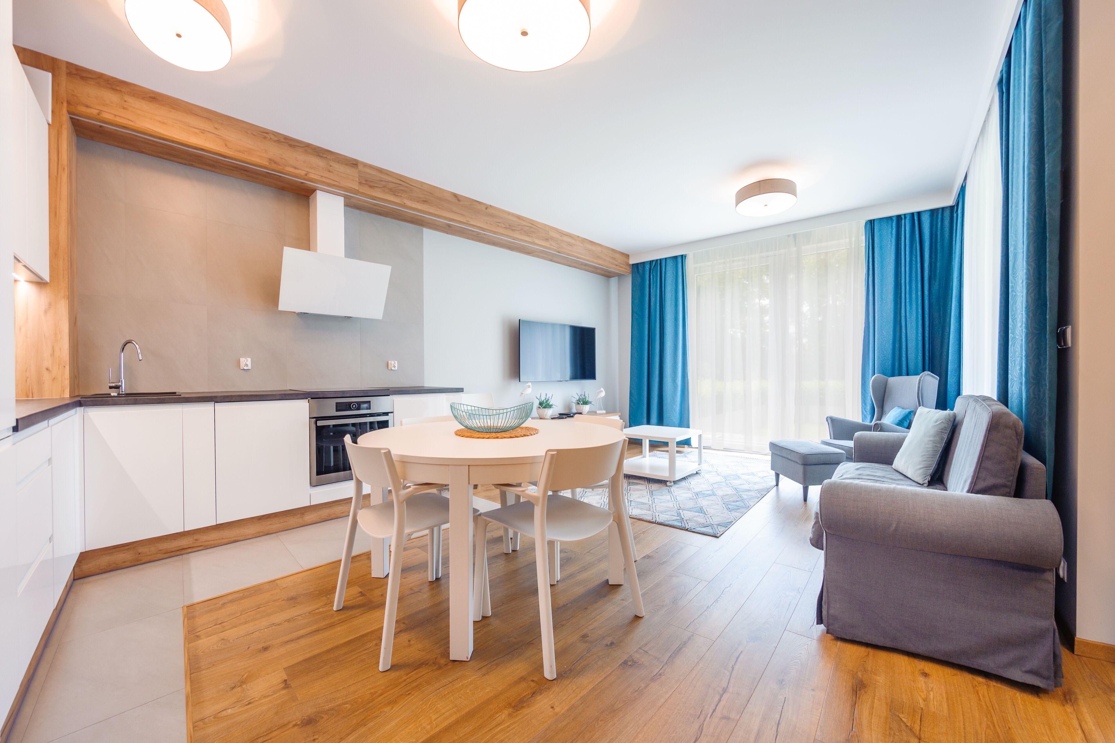A modern living room with a grey sofa, a dining table with chairs, a kitchen area with white cabinets, and a large window with blue curtains.
